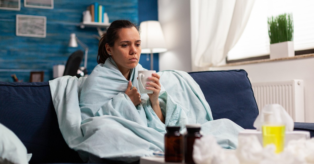 person sitting on a couch surrounded by tissues huddled in a blanket with medications in view.